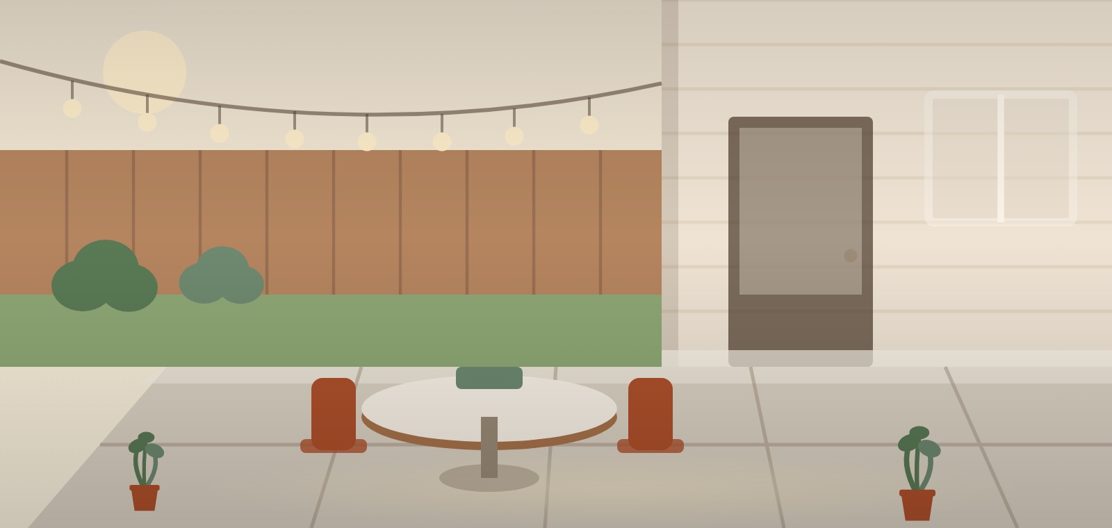 A concrete patio outside a house wall, with a table and chairs, planters and string lights